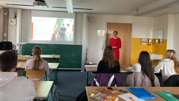 Man sieht von hinten in ein Klassenzimmer, über der Tafel wird eine Präsentation gezeigt, daneben steht eine Frau in einem roten Kleid. Man sieht fünf Schülerinnen und Schüler von hinten, die auf die Präsentation blicken.