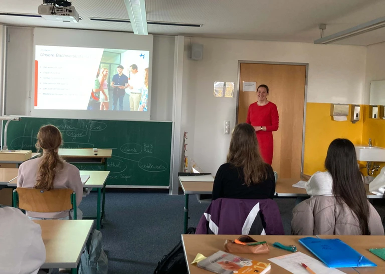 Man sieht von hinten in ein Klassenzimmer, über der Tafel wird eine Präsentation gezeigt, daneben steht eine Frau in einem roten Kleid. Man sieht fünf Schülerinnen und Schüler von hinten, die auf die Präsentation blicken.