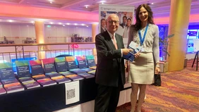 A woman and a man standing in front of a table full of books, smiling into the camera. The man is holding a book in his hands.