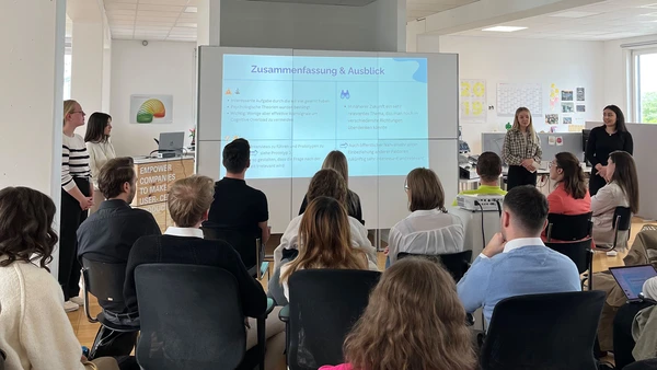 You can see a group of spectators looking at a presentation from behind, and two people speaking in front.