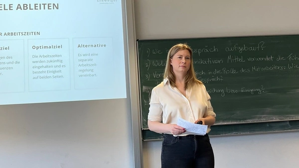 A woman in front of a blackboard, a presentation is shown next to her on the wall.