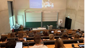 In a lecture hall, in the foreground students from behind, in the background a man giving a lecture.