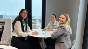 Three female students at a round table smile into the camera.n an einem runden Tisch lächeln in die Kamera.