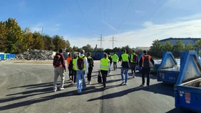 A group of people wearing high-visibility vests from behind next to several blue skip containers.