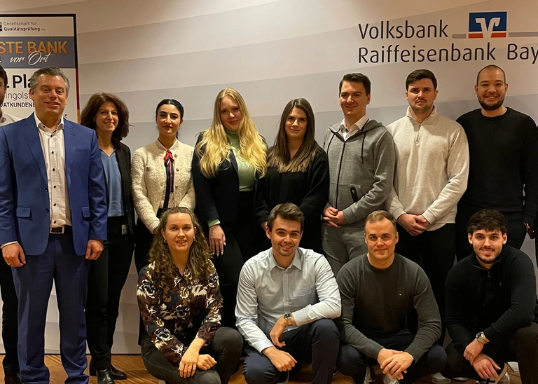 Ein Gruppenfoto mit 14 Personen, im Hintergrund das Logo der VR-Bank Bayern Mitte.