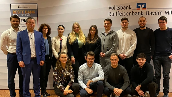 Ein Gruppenfoto mit 14 Personen, im Hintergrund das Logo der VR-Bank Bayern Mitte.