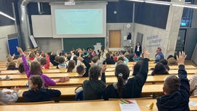 You can see a lecture hall full of children from behind, some of the children come forward, the professor looks into the audience.