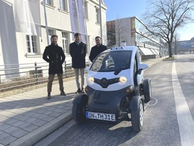 Ein kleines Auto steht an der Straße, im Hintergrund stehen drei Männer