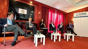 Six people sit next to each other in a red room, a lady in the middle shows various robot models.