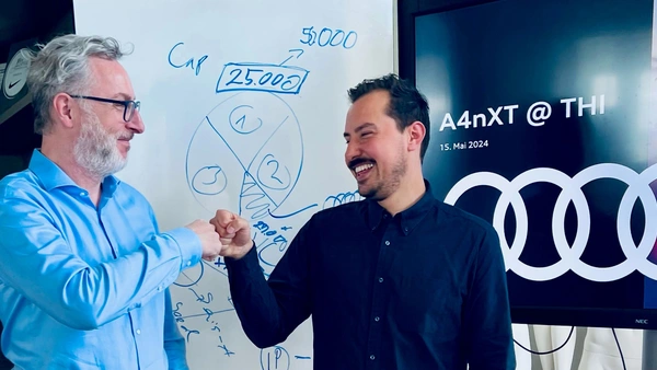 Two men stand opposite each other and laughingly give each other a fist salute, in the background a whiteboard with writing on it and a television on which a presentation is playing.