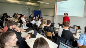 In the lecture hall, you can see the students from the back looking towards the front at a woman giving a presentation.